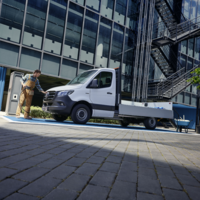Mercedes-Benz eSprinter Pritschenfahrzeug auf Ladefläche vor Bürogebäude – Handwerker nähert sich dem Fahrzeug, Schubkarre daneben