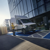 eSprinter mit Kofferaufbau beim Laden an Ladesäule vor Bürogebäude – Fahrer mit Tablet oder Scanner in gelber Jacke